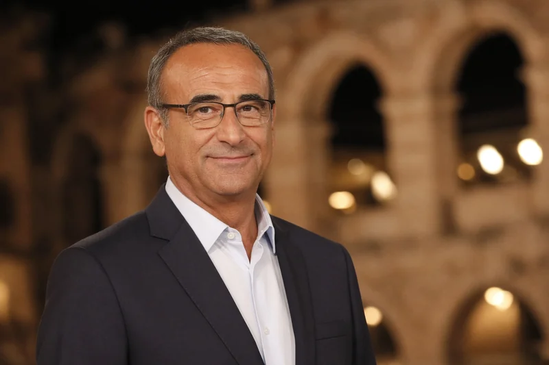 Scena editoriale contestuale generata con IA: Carlo Conti riconoscibile, occhiali e abito scuro, palco notturno ispirato all’Arena di Verona; somiglianza sintetica, non è una fotografia documentaria della serata.