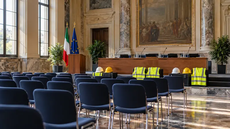 Illustrazione editoriale IA di una sala istituzionale italiana pronta per un convegno, con caschi e giubbotti di sicurezza.