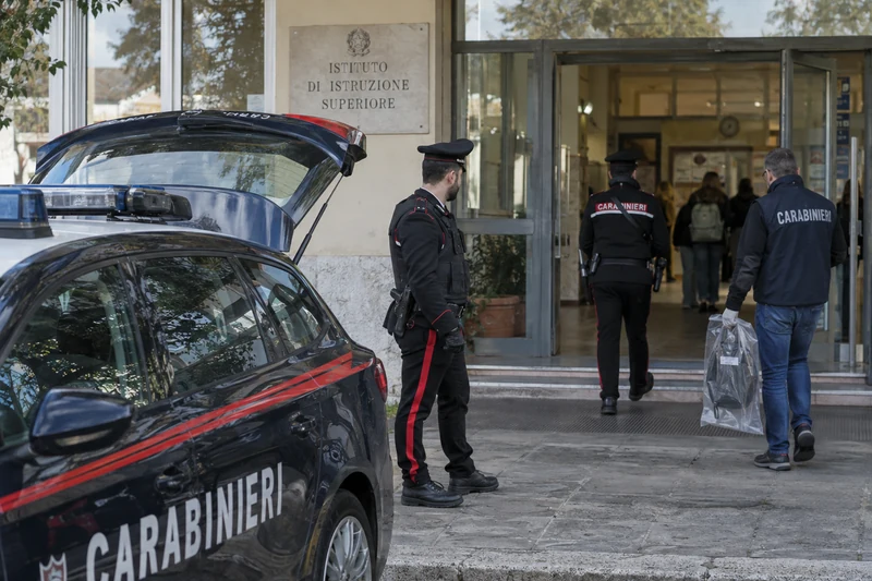 Foto d'archivio di due Carabinieri e di un agente con un sacchetto per prove all'ingresso di un istituto scolastico, con un'auto dei Carabinieri in primo piano.