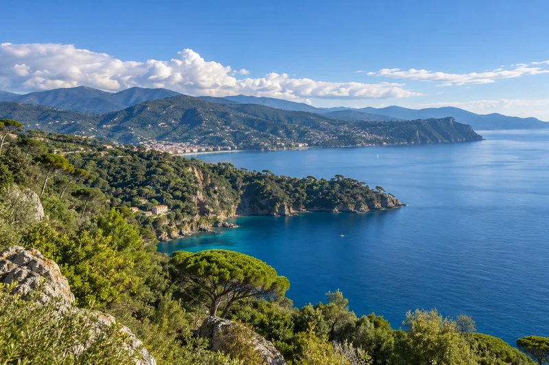 Illustrazione editoriale IA della costa ligure e del Golfo del Tigullio, con sole, mare calmo e nubi sui rilievi; scena non documentaria per il bollettino del 4–11 ottobre 2026.