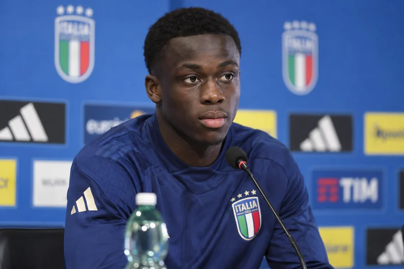 Ritratto editoriale IA di Michael Kayode in una conferenza stampa azzurra; non è una fotografia dell’incontro.