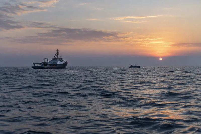 Illustrazione editoriale generata con IA: un'imbarcazione di soccorso e una piccola barca nel Mediterraneo al tramonto; scena simbolica, non documentaria.