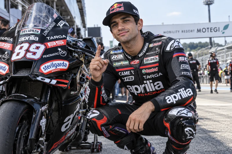 Illustrazione IA di Jorge Martín in tuta Aprilia accanto alla moto nel paddock di Motegi.