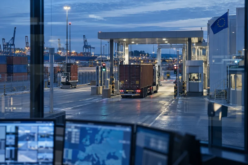 Scena editoriale contestuale generata con IA: terminal merci europeo con varco doganale, camion, container e bandiera UE; non è una fotografia documentaria.