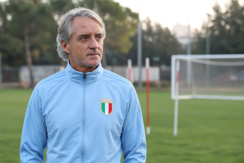 Illustrazione editoriale generata con IA: Roberto Mancini in tuta azzurra della Nazionale sul campo di Coverciano, scena contestuale ordinaria; non è una fotografia documentaria.