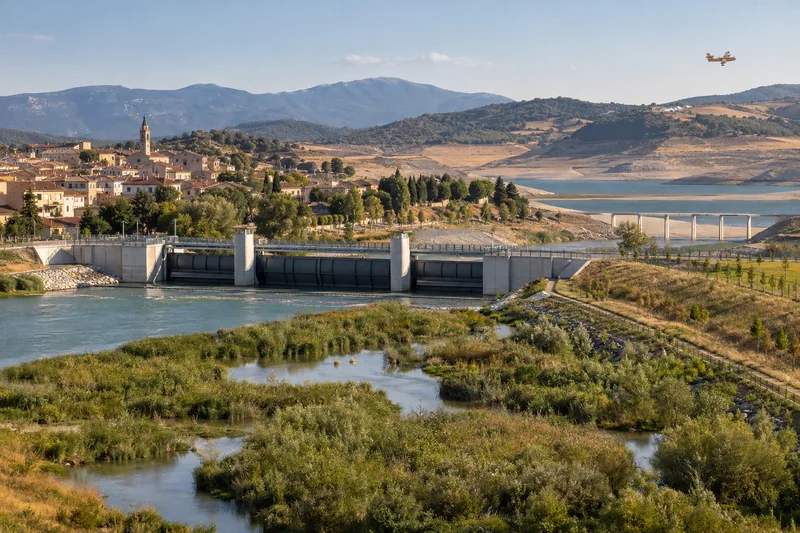 Illustrazione editoriale IA di un centro europeo protetto da una barriera fluviale, tra zone umide ripristinate e territorio colpito dalla siccità; non documentaria.