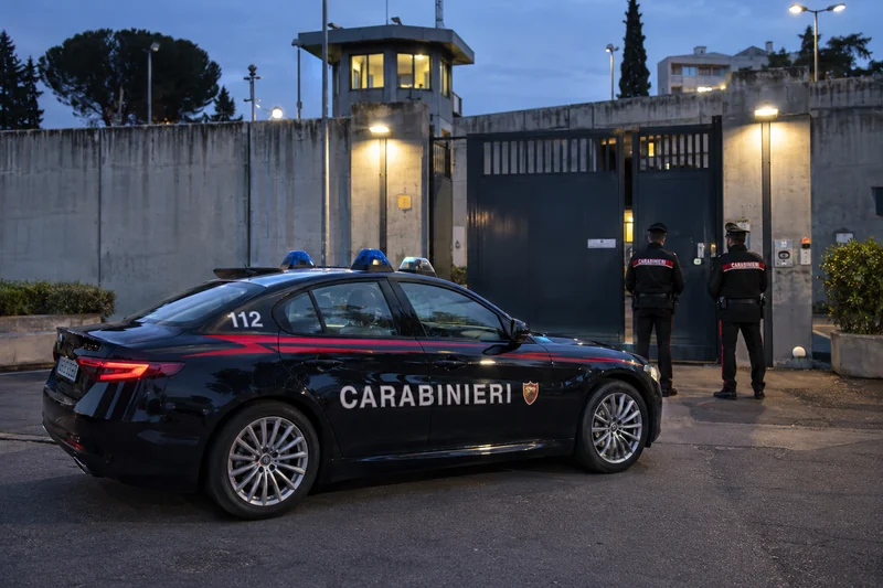 Scena editoriale neutrale generata con IA: auto dei Carabinieri con scritte CARABINIERI e 112 davanti all’ingresso di un istituto penitenziario, senza persone arrestate o vittime visibili; non è una fotografia documentaria.