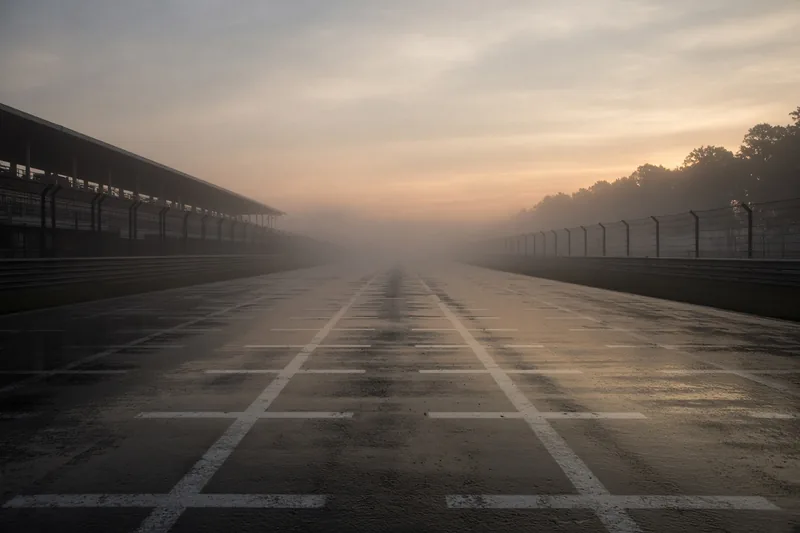 Griglia di partenza vuota all’alba, asfalto bagnato e nebbia; illustrazione editoriale IA, non una fotografia documentaria.